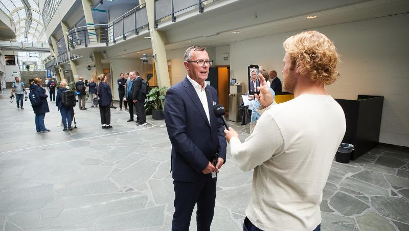 Mann i dress blir intervjuet av en mannlig journalist som holder en mikrofon og filmer med mobiltelefonen